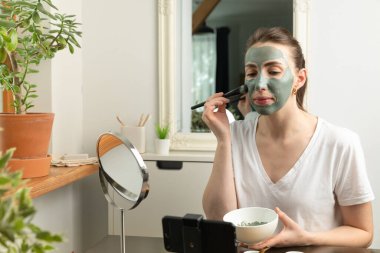 Çevrimiçi cilt bakımı kursuna katılan güzel genç kadınlar akıllı telefonun önüne yeşil kilden bir maske takıyorlar. Güzellik ve cilt bakımı.