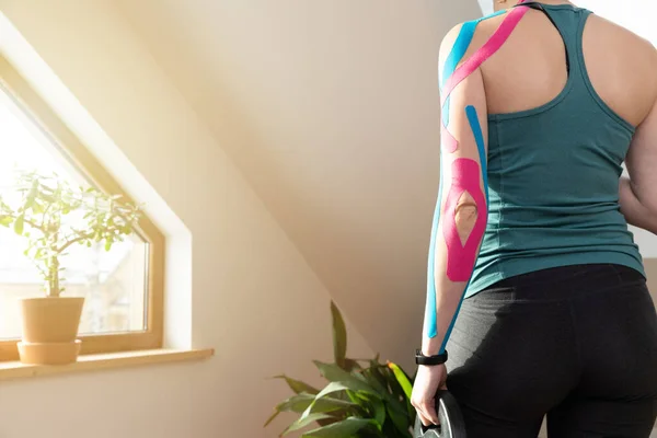 年轻健康的女人站在窗边 手里拿着运动的重物 在她的胳膊上贴上弹性运动学胶带 肌肉治疗 运动和理疗 在家做运动 图库图片