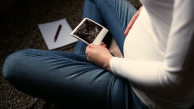 Hamile bir kadın bebek bekliyor, ultrasonu tutuyor ve karnını ovuyor.