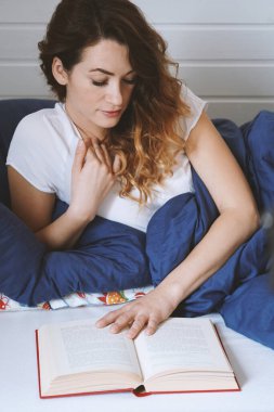 Genç bir kadın yatakta uzanmış kitap okuyor.