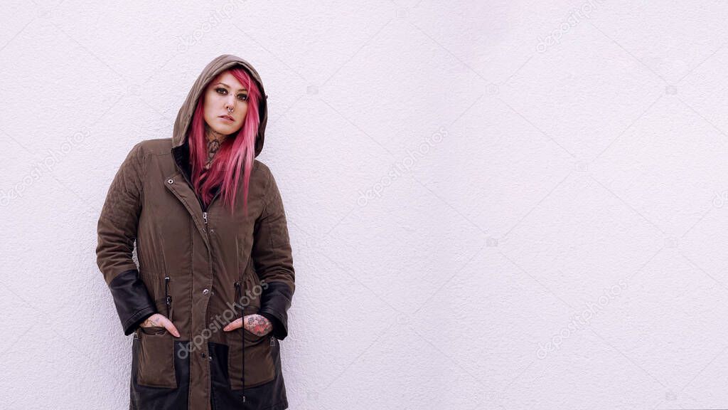 young woman with hooded parka pink hair piercings and tattoos against wall with copy space
