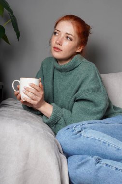 Elinde kupa tutan genç bir kadın, bir kanepede oturuyor, dalgın dalgın dalgın dalgın dalgın dalgın dalgın dalgın dalgın dalgın dalgın bakıyor