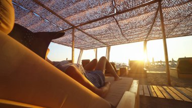 Bronz tenli kadın, kumsalda uzanan şapkalı lüks yatak şezlongunda gün batımında gölgelik altında.