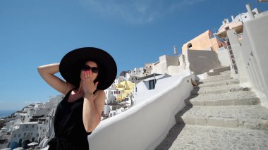 Güneş şapkalı çekici kadın Santorini, Oia merdivenlerinde yürüyor ve kameraya öpücük yolluyor.
