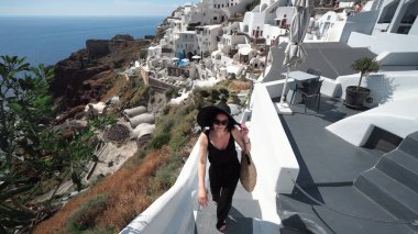 Zarif siyah elbiseli ve güneş şapkalı mutlu manken Santorini, Yunanistan 'da tatilinin tadını çıkarıyor. Otel merdivenlerinde yürümek ve gülümseyen bir kameraya öpücük vermek.