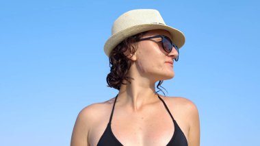 Mutlu, güzel, güler yüzlü, bikinili ve şapkalı bir kız plajda mavi gökyüzüne karşı dans ediyor.