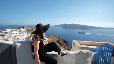 Mutlu gülümseyen siyah elbiseli zarif kadın Santorini 'deki Oia' nın dar kablo kaldırım duvarında oturuşunun keyfini çıkar.