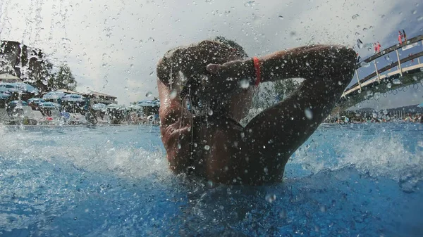 Portrait of beautiful woman relaxing on water jet drops falling down in swimming pool, slow motion