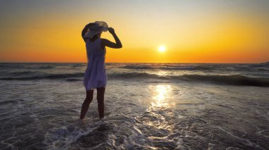 Şapkalı genç bir kadın kumsalda suyun içinde duruyor ve gökyüzüne bakıyor.