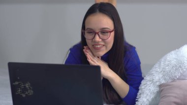 Bilgisayarı kullanan gülümseyen genç bir kadın. İnternette eğleniyor ya da yatakta uzanırken arkadaşlarıyla sohbet ediyor.