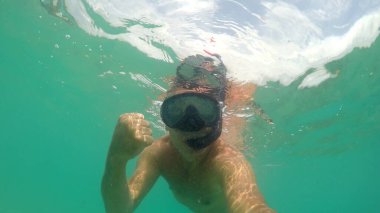 Maskeli şnorkelli adam denize dalıyor ve parmağıyla işaret gösteriyor.