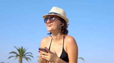 Tropikal sahilde mesajlaşan ve gülen güzel, mutlu genç bir kadın. Hasır şapkalı bir kadın sahilde eğleniyor..
