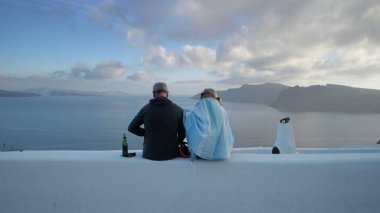 Santorini, Yunanistan - 25 Mayıs 2019: Santorini yaz tatilinde kıdemli çiftler duvarda oturup bira içerek ve çatıların muhteşem panoramik manzarasını seyrederek eğleniyorlar