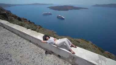 Duvarda uzanmış rahatlamanın keyfini çıkaran bir kadın. Santorini 'deki Vew volkan, kruvazör tekneleri ve kaldera manzaralı.
