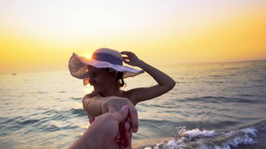 Günbatımında balayında romantik bir çift. Sahilde erkek arkadaşının elini tutarak yürüyen şapkalı, gülümseyen bir kız arkadaş. Yaz tatilinin tadını çıkaran bir çift.