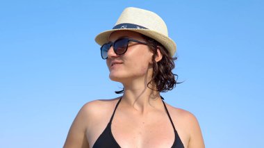 Mutlu, güzel, güler yüzlü, bikinili ve şapkalı bir kız plajda mavi gökyüzüne karşı dans ediyor.