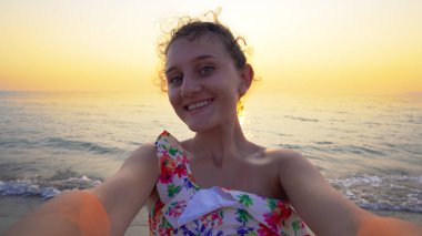 Bikinili güzel bir genç, arka planda yaz mevsiminin günbatımı ile komik selfie suratları yapar.