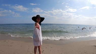 Hasır şapkalı mutlu kadın sevgilisiyle el ele tutuşuyor ve kumsalda eğleniyor, ağır çekimde.