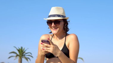 Tropikal sahilde mesajlaşan ve gülen güzel, mutlu genç bir kadın. Hasır şapkalı bir kadın sahilde eğleniyor..