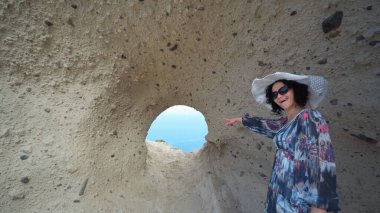 Uzun elbiseli ve şapkalı kadın, Yunanistan 'ın Santorini kentinin bir simgesi olan Santorini' nin Kalbi 'nden Santorini manzarasının keyfini çıkarıyor