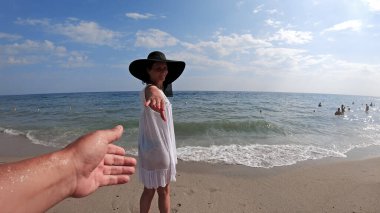 Hasır şapkalı mutlu kadın sevgilisiyle el ele tutuşuyor ve kumsalda eğleniyor, ağır çekimde.