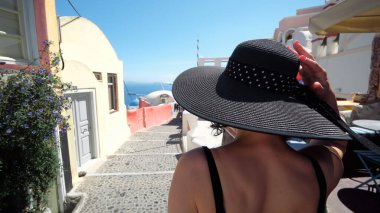 Oia, Santorini, Yunanistan 'da seyahat tatilinde şapkalı zarif bir turist kadın. Oia 'da dar bir sokakta yürüyen mutlu genç kadın Yunan adasını ziyaret ediyor..