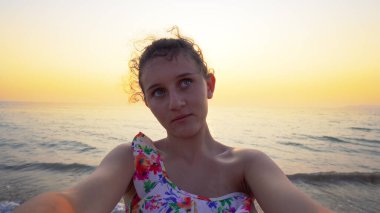 Bikinili güzel bir genç, arka planda yaz mevsiminin günbatımı ile komik selfie suratları yapar.