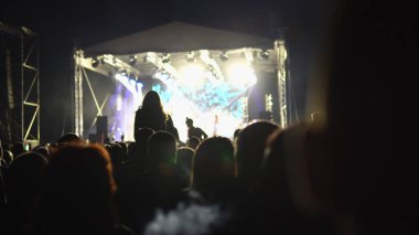 Sahne ışıklarıyla aydınlatılmış büyük bir sahnenin önünde insan siluetlerinin alkışladığı bir konser.