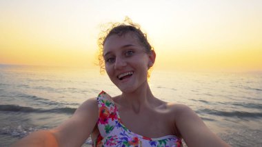 Bikinili güzel bir genç, arka planda yaz mevsiminin günbatımı ile komik selfie suratları yapar.