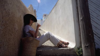 Santorini 'de dar bir caddenin merdivenlerinde oturan ve bahar çiçeklerini koklayan gezgin kadın Santorini' de dar bir caddenin merdivenlerinde oturmuş bahar çiçeği kokluyor.