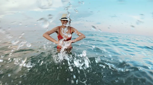 Slow motion of a beautiful girl with straw hat being into the sea and splashing towards the camera