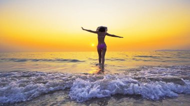 Bikinili ve şapkalı seksi genç gün batımında deniz suyundan çıkıyor, yaz tatili konsepti.