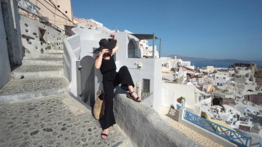 Siyah elbiseli zarif kadın Santorini 'deki Oia' nın dar kablo kaldırım duvarında oturmasının tadını çıkarıyor.
