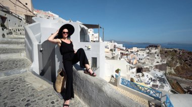 Mutlu gülümseyen siyah elbiseli zarif kadın Santorini 'deki Oia' nın dar kablo kaldırım duvarında oturuşunun keyfini çıkar.