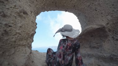 Uzun elbiseli ve şapkalı kadın, Yunanistan 'ın Santorini kentinin bir simgesi olan Santorini' nin Kalbi 'nden Santorini manzarasının keyfini çıkarıyor