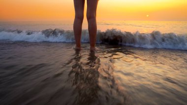 Sahilde gün batımında kadın ayaklarına su sıçrattı. Huzurlu ve rahatlatıcı okyanus suyu sahnesi güzel sahilde ayaklara sıçrıyor.