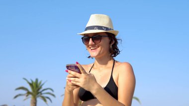Tropikal sahilde mesajlaşan ve gülen güzel, mutlu genç bir kadın. Hasır şapkalı bir kadın sahilde eğleniyor..