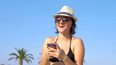 Tropikal sahilde mesajlaşan ve gülen güzel, mutlu genç bir kadın. Hasır şapkalı bir kadın sahilde eğleniyor..