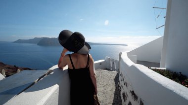Romantik Santorini tatili. Siyah giyinmiş zarif bir kadın, Santorini manzarasının keyfini çıkarıyor. Deniz seviyesinin üzerinde, dar bir sokakta duruyor.
