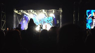 Sahne ışıklarıyla aydınlatılmış büyük bir sahnenin önünde insan siluetlerinin alkışladığı bir konser.