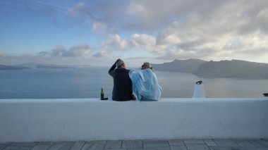 Santorini, Yunanistan - 25 Mayıs 2019: Santorini yaz tatilinde kıdemli çiftler duvarda oturup bira içerek ve çatıların muhteşem panoramik manzarasını seyrederek eğleniyorlar