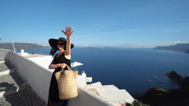 Siyahlı zarif esmer kadın duvarın yanında durmuş Oia 'nın muhteşem Santorini panoramik manzarasının keyfini çıkarıyor.