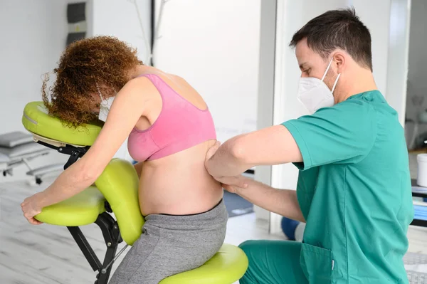 Colpo di giovane fisioterapista che massaggia la schiena di donna incinta. Uomo con maschera protettiva durante la nuova normale. — Foto Stock