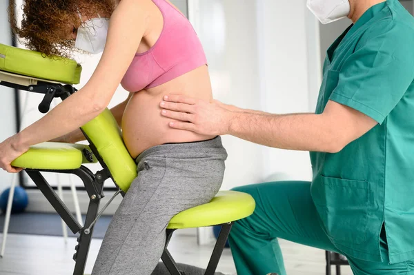 Colpo di giovane fisioterapista che massaggia la schiena di donna incinta. Uomo con maschera protettiva durante la nuova normale. — Foto Stock