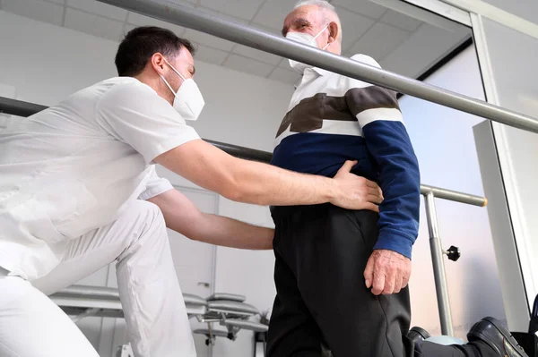 Fisioterapista che aiuta il paziente anziano a camminare tra le sbarre parallele. — Foto Stock