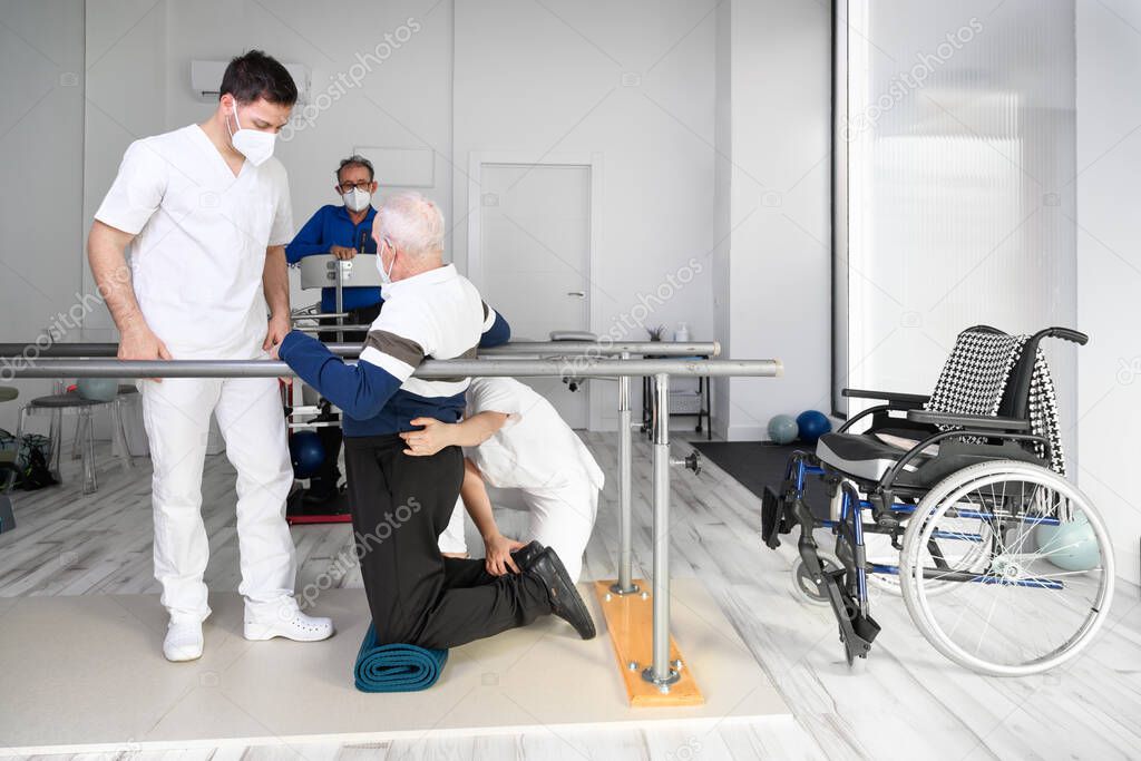 Physiotherapists helping disabled Caucasian senior man to walk with parallel bars in rehabilitation center. 