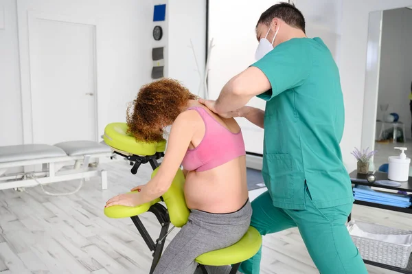 拍年轻理疗师按摩孕妇背。新常态下戴防护口罩的男子. — 图库照片