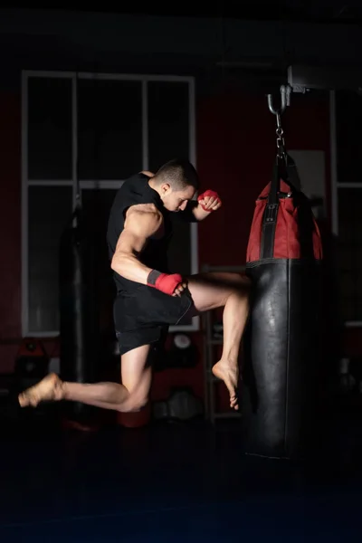 Kickboxing combattente Esecuzione salto aria calci con ginocchio sul sacco da boxe. caucasico uomo pratica arti marziali formazione a urbano palestra. — Foto Stock