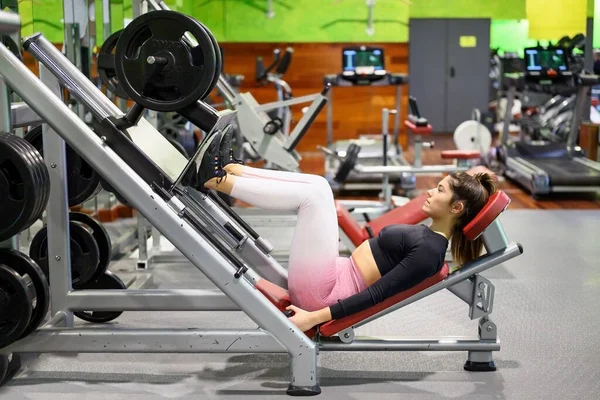 Vue latérale de la forme jeune sportive fléchissant les muscles de ses jambes sur la machine d'exercice à la salle de gym. — Photo
