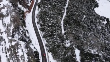 Karlı buz yolunda giden kırmızı bir arabanın hava aracı kışın yerel manzaraları keşfediyor, güzel kozalaklı ormanlarla çevrili bölgede hareket eden otomobilin kuş bakışı görüntüsü.